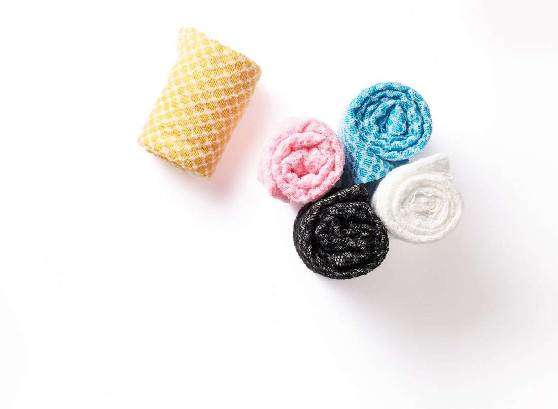 Five rolled Sima Japanese exfoliating towels in yellow, pink, blue, black and white on a white background