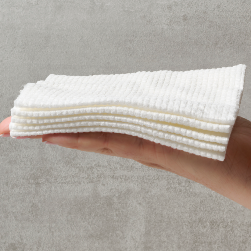 Hand holding a fanned-out stack of white Sima disposable face towels against a gray background