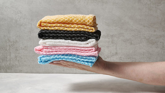 Hand holding a stack of folded Sima Japanese exfoliating towels in yellow, black, white, pink and blue against a gray wall