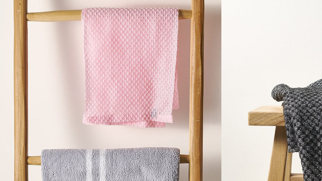 Pink Sima Japanese exfoliating towel hung flat to dry beside a black towel draped over a wooden stool