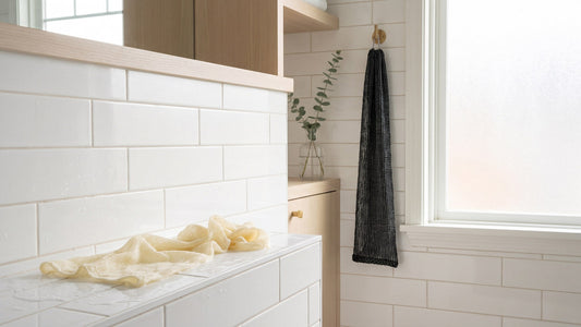 Exfoliating towel hanging from a hook on white bathroom tiles beside a wet cream cloth on the ledge