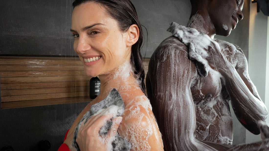 Smiling woman in the shower gently washing her shoulder and upper arm with a dark exfoliating towel