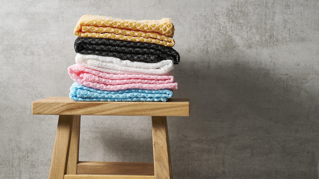 Stack of folded Sima Japanese exfoliating towels in yellow, black, white, pink and blue on a wooden stool against a gray wall